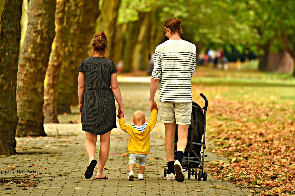 Eltern mit ihrem Kleinkind zwischen sich an den Händen gehen unter herbstlichen Bäumen entlang
